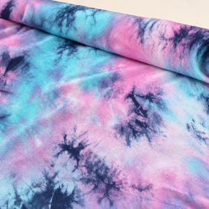 May include: A bolt of fabric featuring a tie-dye pattern with blended shades of pink, light blue, and dark navy blue. The design consists of soft, cloud-like color transitions interspersed with darker, starburst-shaped ink splatters throughout.