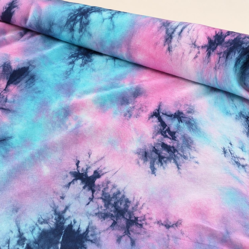 May include: A bolt of fabric featuring a tie-dye pattern with blended shades of pink, light blue, and dark navy blue. The design consists of soft, cloud-like color transitions interspersed with darker, starburst-shaped ink splatters throughout.