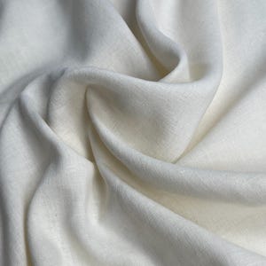 Pode incluir: Close-up de um tecido de linho creme com dobras suaves e textura sutil. O tecido parece leve e fluido, com um tom natural ligeiramente esbranquiçado. A imagem destaca a queda e a textura do material.