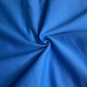 Pode incluir: Close-up de um tecido azul vibrante com uma textura macia. O tecido é reunido em espiral, criando dobras e sombras que realçam sua textura. A cor é um azul brilhante e sólido, adequado para roupas ou decoração de casa.