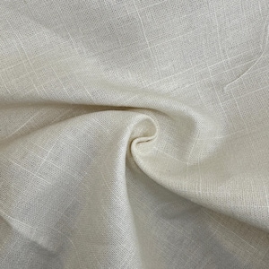 Puede incluir: Primer plano de tela de lino blanquecina con un tejido texturizado. La tela está doblada y drapeada, creando sombras suaves y resaltando la textura natural. El material parece ligero y transpirable, adecuado para ropa o decoración del hogar.