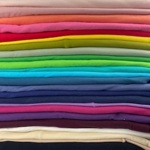 Puede incluir: Una pila de tela doblada en una variedad de colores, desde rosa claro hasta morado oscuro. La tela parece ser suave, posiblemente algodón o un textil similar. Los colores están dispuestos en un degradado, creando una exhibición visualmente atractiva.