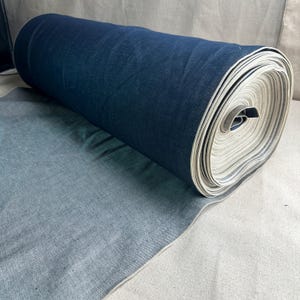 Puede incluir: Un rollo de tela vaquera índigo oscuro enrollada. La tela está desenrollada, revelando un tono azul más claro. Los bordes de la tela son visibles, mostrando la textura y el tejido del material. La tela es adecuada para ropa y decoración del hogar.