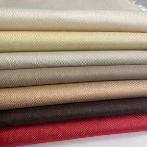 Puede incluir: Una pila de muestras de tela en varios colores neutros, incluyendo crema, beige, marrón y rojo. La tela tiene un tejido texturizado, y los bordes están cuidadosamente cortados. La imagen muestra una gama de opciones textiles.