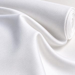 May include: A roll of solid white fabric featuring a distinct diagonal twill weave pattern. The material is tightly wound on one end and cascades into soft, flowing folds, highlighting the consistent texture and bright white color throughout.