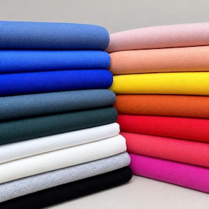 May include: A stack of folded fabric pieces in a variety of solid colors including shades of blue, gray, black, white, pink, red, orange, and yellow. Each piece of fabric features a consistent, fine-textured weave and is neatly layered.