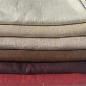 Puede incluir: Una pila de tela de lino doblada en varios colores neutros. Los colores incluyen blanco roto, beige, marrón claro, marrón, burdeos y rojo. La tela tiene un aspecto texturizado, lo que sugiere una fibra natural.