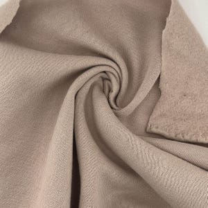 Pode incluir: Close-up de um pedaço de tecido dobrado em uma cor marrom claro neutra. O tecido tem uma textura canelada de um lado e uma textura mais macia e feltrada do outro. O material parece ser um tecido de malha ou lã polar.