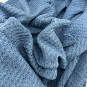 Pode incluir: Close-up de um tecido de malha waffle azul macio. O tecido é dobrado e drapeado, mostrando a textura e o padrão da malha. A cor é um azul suave e empoeirado, e o tecido parece leve e confortável.