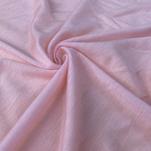 Pode incluir: Close-up de um redemoinho de tecido rosa claro e macio. O tecido tem um brilho sutil e parece leve, com dobras e vincos visíveis. A cor é um pastel delicado, sugerindo uma textura suave e arejada.