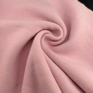 Pode incluir: Close-up de um redemoinho de tecido rosa macio. O material parece ser malha ou lã polar, com uma textura suave e variações sutis de tom. O tecido está bem dobrado, criando um padrão dinâmico e rodopiante.