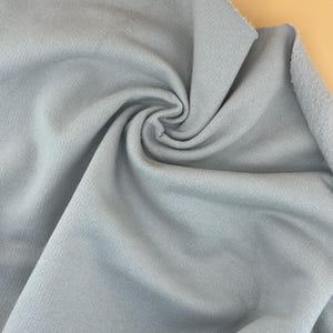 Pode incluir: Close-up de um tecido azul claro com uma aparência macia e texturizada. O tecido é rodopiado, criando um padrão em espiral. O material parece ser um tecido de malha ou têxtil semelhante, adequado para roupas ou decoração de casa.
