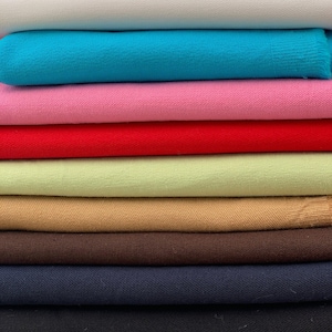 Puede incluir: Una pila de tela doblada en varios colores. Los colores incluyen blanco, turquesa, rosa, rojo, verde claro, beige, marrón, azul marino y negro. La tela parece ser un material de punto o tejido.