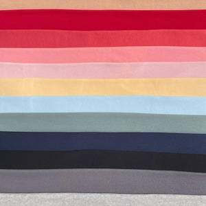 Puede incluir: Una paleta de colores de muestras de tela en varios tonos. Las muestras están dispuestas horizontalmente, mostrando una gama de colores que van desde el marrón y el beige hasta el rojo, rosa, amarillo, azul y gris. Las telas parecen suaves y texturizadas.