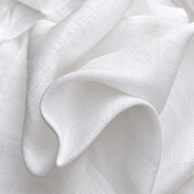 May include: A piece of white fabric with a visible woven texture and a natural, slightly irregular weave pattern. The material is draped in soft, flowing folds, highlighting the matte finish and the crisp, clean appearance of the white textile.