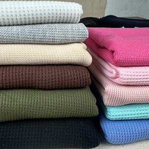 Puede incluir: Una pila de telas de punto de gofre dobladas en varios colores. Los colores incluyen blanco, gris, beige, marrón, verde oliva, negro, rosa, rosa claro, turquesa y azul. La tela tiene un patrón texturizado en forma de rejilla.