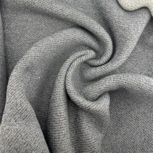 Puede incluir: Primer plano de tela gris con un tejido texturizado. El material está doblado y drapeado, creando curvas y sombras suaves. La tela parece ser de peso medio, adecuada para proyectos de ropa o decoración del hogar.