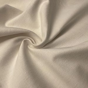 Pode incluir: Close-up de tecido branco sujo com uma superfície texturizada e canelada. O tecido é dobrado e drapeado, criando sombras suaves e destacando a textura do material. A cor é um branco quente e cremoso.
