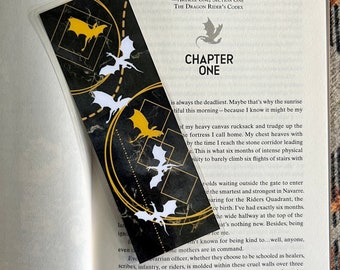 May include: A black bookmark with a gold border and a repeating pattern of white dragons. The bookmark has the text "CHAPTER ONE" and a quote from "The Dragon Rider's Codex".