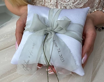 May include: White satin pillow with a light green ribbon and bow. The pillow has the text "Stephen + Aimee" and "June 24, 2023" printed on it.