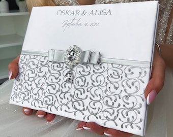 May include: White wedding guest book with silver glitter swirls and a silver ribbon with a rhinestone bow and pendant. The cover reads "OSKAR & ALISA September 16, 2026".