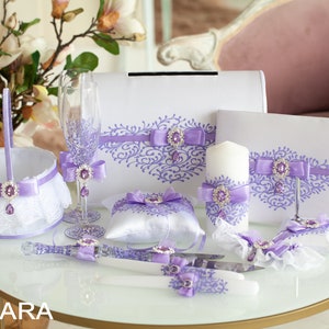 Op de afbeelding: Een huwelijksceremonie set met lavendel accenten. De set bevat een ringkussen, een taartmes, een kaars, een mand, twee champagne glazen en twee gastenboeken.