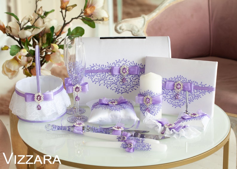Op de afbeelding: Een huwelijksceremonie set met lavendel accenten. De set bevat een ringkussen, een taartmes, een kaars, een mand, twee champagne glazen en twee gastenboeken.