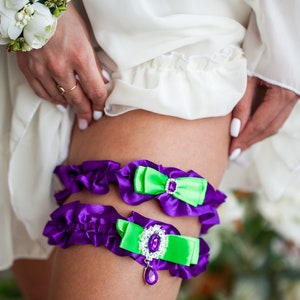 May include: Purple and green garter with a jeweled embellishment and a bow. The garter is made of satin and lace.