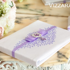 Op de afbeelding: Wit bruiloftsgastenboek met een lavendelkleurig lint en een juweelversiering. Het boek heeft een decoratief bloemmotief dat op de omslag is gedrukt. De tekst "VIZZARA" is zichtbaar op de achtergrond.
