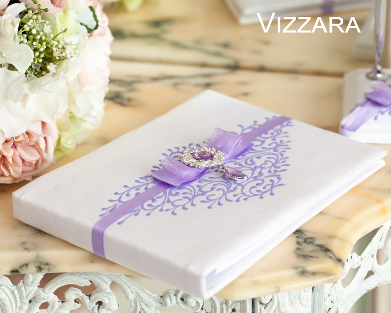Op de afbeelding: Wit bruiloftsgastenboek met een lavendelkleurig lint en een juweelversiering. Het boek heeft een decoratief bloemmotief dat op de omslag is gedrukt. De tekst "VIZZARA" is zichtbaar op de achtergrond.