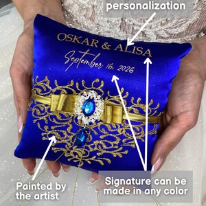 May include: Blue satin ring pillow with gold embroidery and a blue jeweled embellishment. The pillow is personalized with the names "OSKAR & ALISA" and the date "September 16, 2026".