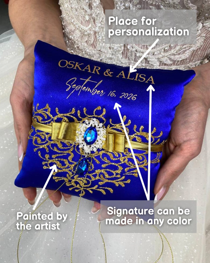 May include: Blue satin ring pillow with gold embroidery and a blue jeweled embellishment. The pillow is personalized with the names "OSKAR & ALISA" and the date "September 16, 2026".