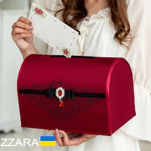 May include: A red satin wedding card box with a black ribbon and a jeweled embellishment. The box is decorated with a black floral design.