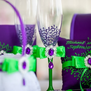 May include: Two champagne flutes with purple and green accents. The flutes are decorated with a purple floral design and have green bows with a jeweled center. The flutes are sitting on a purple and green pillow with the text "Michael and September" on it.