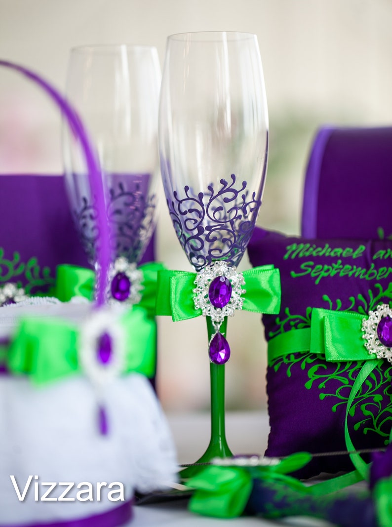May include: Two champagne flutes with purple and green accents. The flutes are decorated with a purple floral design and have green bows with a jeweled center. The flutes are sitting on a purple and green pillow with the text "Michael and September" on it.