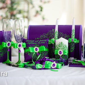 May include: Purple and green wedding ceremony set with two champagne flutes, a ring pillow, two unity candles, a guest book, a cake knife and server, and a flower girl basket. The set is decorated with green and purple ribbons and rhinestones.