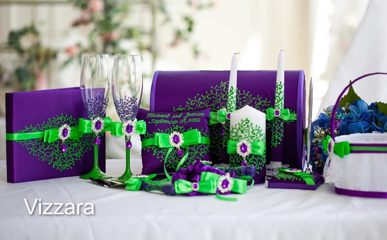 May include: Purple and green wedding ceremony set with two champagne flutes, a ring pillow, two unity candles, a guest book, a cake knife and server, and a flower girl basket. The set is decorated with green and purple ribbons and rhinestones.