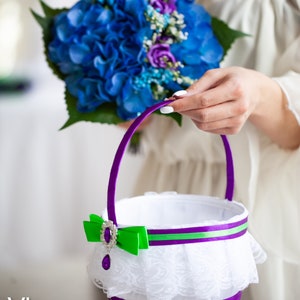 May include: A white flower girl basket with a purple satin ribbon, green and purple ribbon, and a white lace trim. The basket has a purple jewel embellishment.