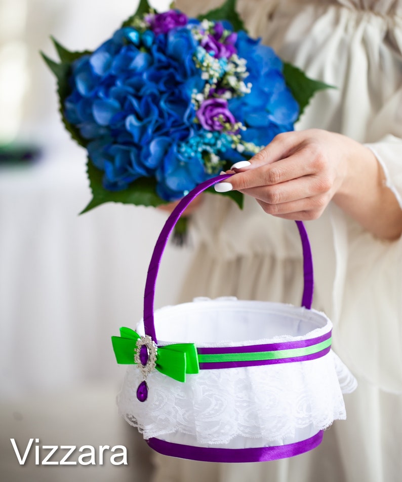 May include: A white flower girl basket with a purple satin ribbon, green and purple ribbon, and a white lace trim. The basket has a purple jewel embellishment.