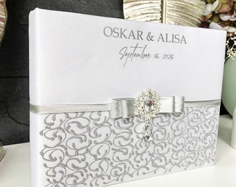 May include: White wedding guest book with a silver glitter design and a silver ribbon with a rhinestone embellishment. The book is personalized with the names "OSKAR & ALISA" and the date "September 16, 2026".