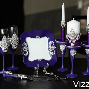 May include: Purple wedding cake serving set with two champagne flutes, a cake knife, a plate, and two candle holders with white candles. The set is decorated with rhinestones.