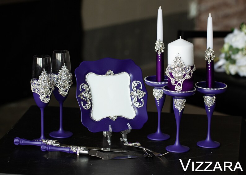 May include: Purple wedding cake serving set with two champagne flutes, a cake knife, a plate, and two candle holders with white candles. The set is decorated with rhinestones.