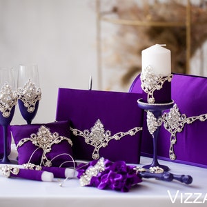 May include: Purple wedding ceremony set with a candle, two champagne flutes, a ring pillow, a box, a basket, and two unity candles. The set is decorated with rhinestones.