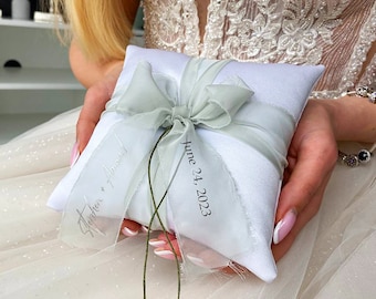 May include: White satin ring pillow with a sage green ribbon and bow. The pillow is embroidered with the text "Stephen + April" and "June 24, 2023".