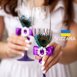 May include: Two clear champagne flutes with purple stems. Each glass is decorated with a purple satin bow, a jewel, and peacock feathers. The glasses are held by a person wearing a white dress. The brand name "Vizzara" is visible.