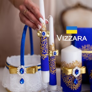 Puede incluir: Dos velas blancas decoradas con cinta azul y dorada y un diseño estampado azul y dorado. Las velas están en una cesta blanca con una cinta azul y un lazo dorado. La bandera de Ucrania está en el fondo.