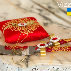 May include: A red satin pillow with gold embroidery and a jeweled bow, along with two silver cake knives with red and gold handles. The knives are tied together with a gold ribbon. The text "VIZZARA" is visible in the upper right corner of the image.