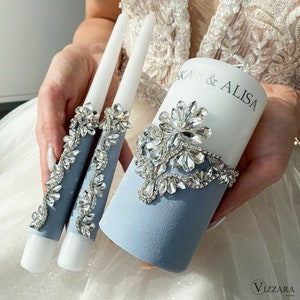Puede incluir: Un conjunto de tres velas de boda blancas con bases de terciopelo azul y adornos de pedrería plateada. La vela central es más alta y tiene los nombres "KAR & ALISA" escritos en ella.
