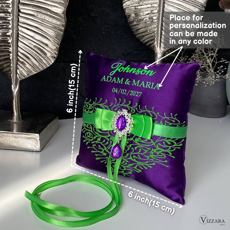 May include: A purple satin ring pillow with green and purple embroidery. The pillow is 6 inches square and has a green satin ribbon with a jeweled brooch. The pillow is personalized with the text "Johnson ADAM & MARIA 04/02/2027".