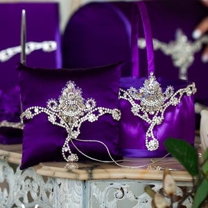 May include: A purple satin ring pillow and flower girl basket with silver rhinestone embellishments. The pillow and basket are decorated with a crown-shaped design.
